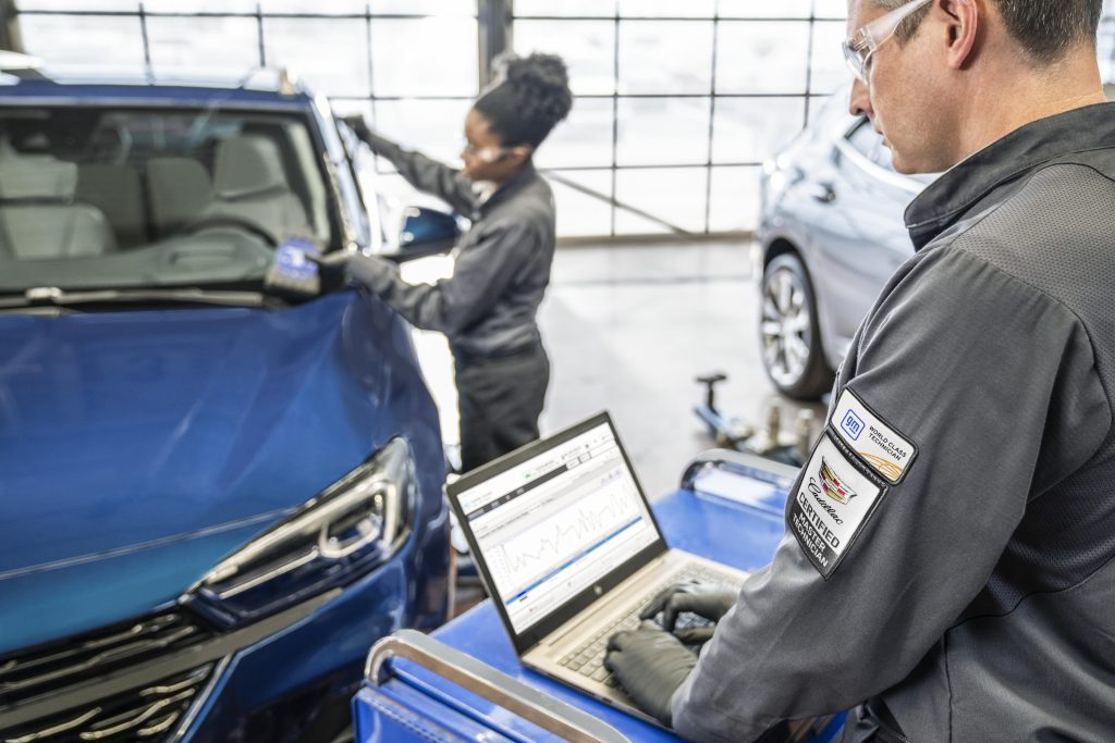 Two technicians working on a vehicle, with one using a laptop for diagnostics 
